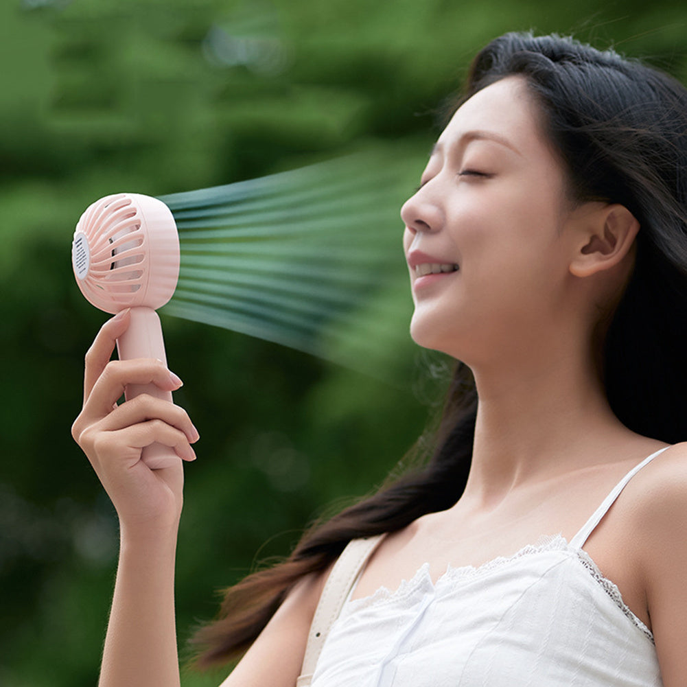A woman in a white sleeveless top enjoys the refreshing breeze from the Glacient Semiconducting Handheld Fan by CJDropshipping, looking content and relaxed outdoors with lush green foliage in the background.