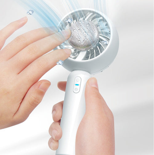 A person holds the CJDropshipping Glacient Semiconducting Handheld Fan, a white USB-rechargeable portable cooling device, with one hand while touching its silver center as blue airflow and water droplets blow from it.