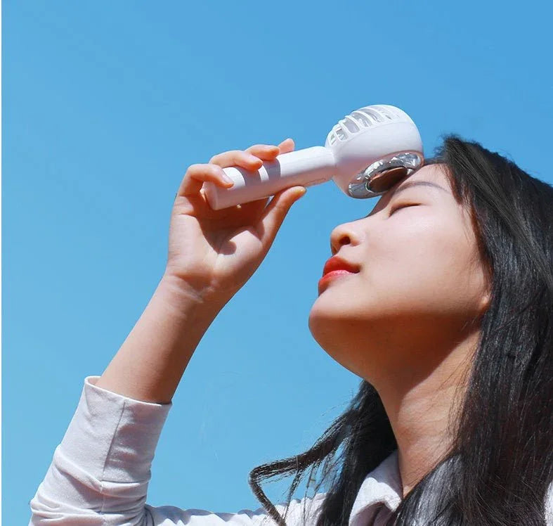 A woman uses the CJDropshipping Glacient Semiconducting Handheld Fan outdoors under a clear blue sky, enjoying instant portable cooling as she holds the small white fan to her forehead.