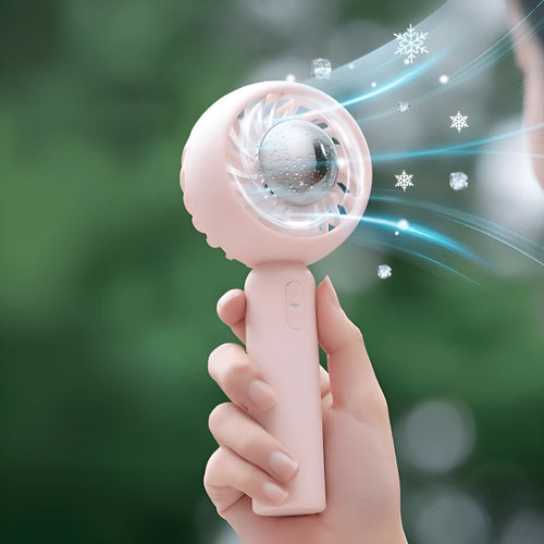 A hand holds the Glacient Semiconducting Handheld Fan by CJDropshipping, a small pink USB rechargeable fan blowing cool air and snowflake icons, evoking portable refreshment against a blurred green backdrop.